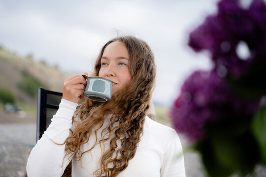 A curly-haired woman enjoys a warm drink by the seaside, the wind blowing through her hair. She sits in a chair and enjoys cozy coastal relaxation in the daytime.