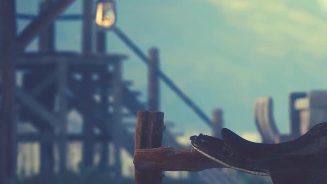 A saddle rests on a wooden stand in front of a rustic building. Wooden steps lead up to the structure. Soft light filters through the trees, creating a serene atmosphere.