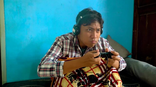 Young Gamer Wearing Headphones Playing Video Game With Controller On Bed Against Blue Wall Background