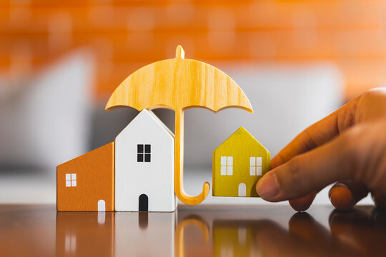 Hand protecting wooden house models under a wooden umbrella. Concept of real estate insurance, home safety, property loan security, and asset protection plan.