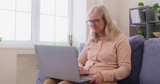 Serious old woman focused at laptop screen, typing email and surfing information using home internet and sitting on sofa at home, communicating online. Old people and technology concept