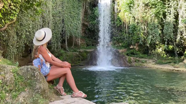 Woman traveler with straw hat sitting by tropical waterfall and emerald natural pool, Spain, Frias