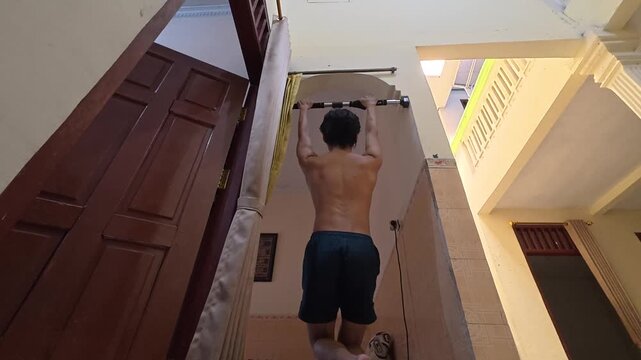 Back view of a shirtless man doing scapula pull ups on a doorway bar at home, strength training