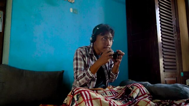 Asian Man Wearing Headphones Focused On Playing Video Games In A Blue Room