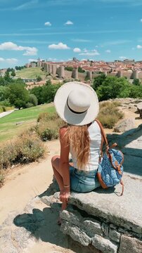 Back view of woman traveler with straw hat and backpack sitting on stone viewpoint looking at Avila medieval walls in Spain- Vertical video