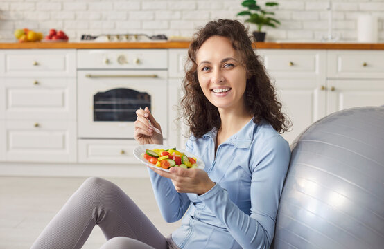 Portrait of a young sporty slim woman wearing sportswear holding plate with healthy food of fresh vegetables sitting on floor in kitchen at home after sport workout. Healthy nutrition concept.