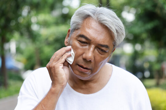 Tired and uncomfortable old asian senior man wiping his face with refreshing cold wet towel, trying to relieve from heavy sweating and outdoor summer heat wave