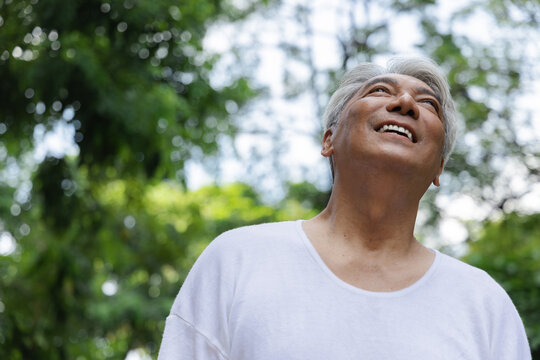 Relaxed happy refreshing old retired senior Asian man looking up and showing smiling expression in urban park, good summer, summer wellness, sunlight exposure concept image