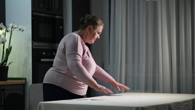 woman covering a dining table with a tablecloth in the evening. Cozy home preparation scene focused on household routine and table setting.