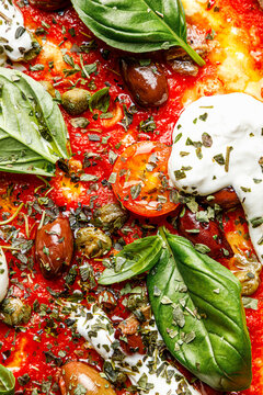 Close-up texture of a pizza with burrata, basil, tomatoes, and olives.