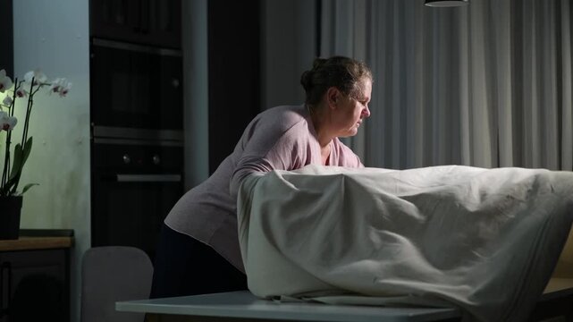 woman covering a dining table with a tablecloth in the evening. Cozy home preparation scene focused on household routine and table setting.