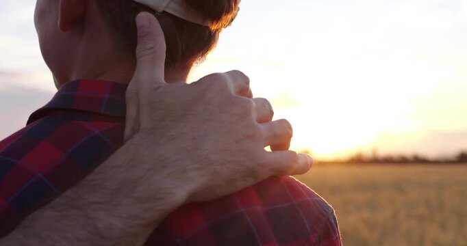 Father gently embraces son while walking through golden wheat field at sunset. Close-up captures protection, trust, support and lasting family bond filled with warmth, care and quiet confidence.