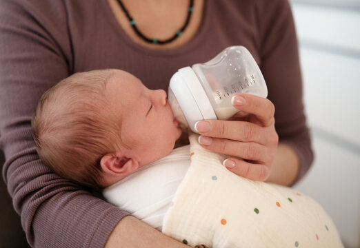 Mom Holds Wrapped Baby While Feeding From Bottle In Bright Room. Relaxed And Nurturing Moment.