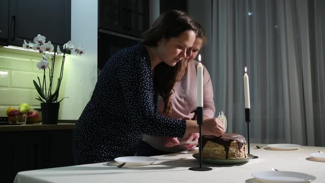 Two women place a birthday cake with number 60 candles on the table and add decorative candles, preparing a milestone celebration at home.