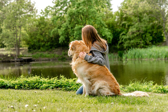 Teen Girl Sitting By Lake With Golden Retriever View From Behind
