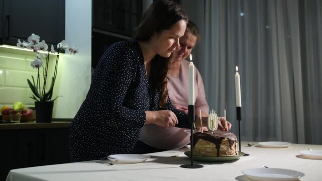 Two women place a birthday cake with number 60 candles on the table and add decorative candles, preparing a milestone celebration at home.