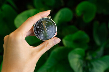 Hand holding a magnetic compass on a blurred green leaves background. Concept of business direction, strategic planning, travel navigation, sustainability, and choosing the right path. © marinasokolchik