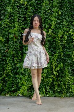 Young woman in floral dress standing barefoot against green leafy wall outdoors