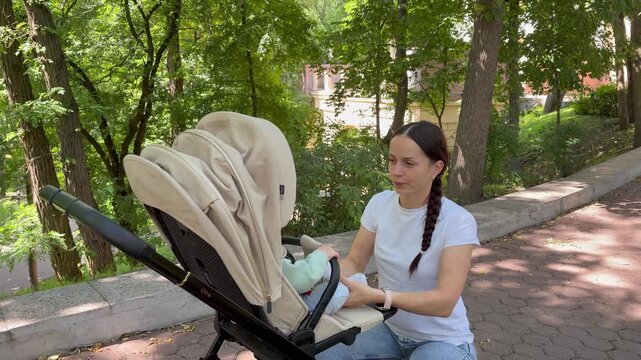 Young mother kneeling beside a beige stroller interacting with her baby in a sunny park. a warm and peaceful atmosphere of family leisure