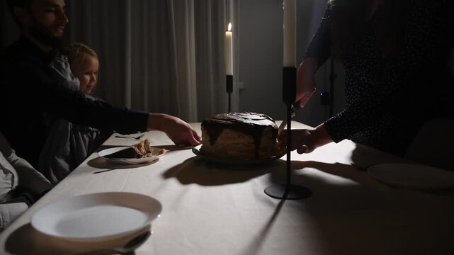 Family gathers around the table in the evening and begins cutting a birthday cake by candlelight, sharing a warm moment together during the celebration.