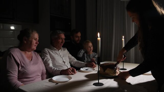 Family gathers around the table in the evening and begins cutting a birthday cake by candlelight, sharing a warm moment together during the celebration.