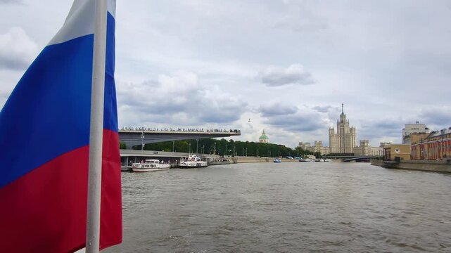 russian flag waves in foreground with floating bridge and stalinist skyscraper view from moscow river cruise. scenic boat tour showcases zaryadye park modern architecture and iconic soviet era tower
