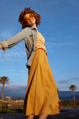 Fototapeta premium Smiling woman in denim jacket and striped top twirls outdoors under bright sky, flowing orange skirt. Lifestyle, outdoor photography, carefree mood and expressive movement.