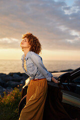 Fototapeta premium A young woman with curly hair leans against a car by the ocean at sunset, posing for a reflective moment.