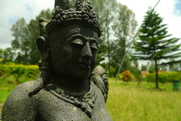 Obraz premium Traditional stone statue of a Balinese deity with intricate carvings and weathered texture captured in a soft natural light outdoors showing serene facial features and an ornate headpiece from close