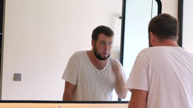 Young man using an electric beard trimmer while standing in front of a mirror. Daily grooming routine, facial hair care, personal hygiene and morning self-care at home.