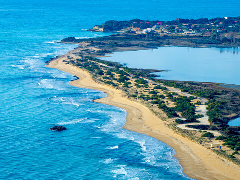 Aerial telephoto view of Halikounas beach curve separating Lake Korission from Ionian Sea in Corfu Greece, featuring sand dunes, green vegetation, and distant houses on a sunny summer day