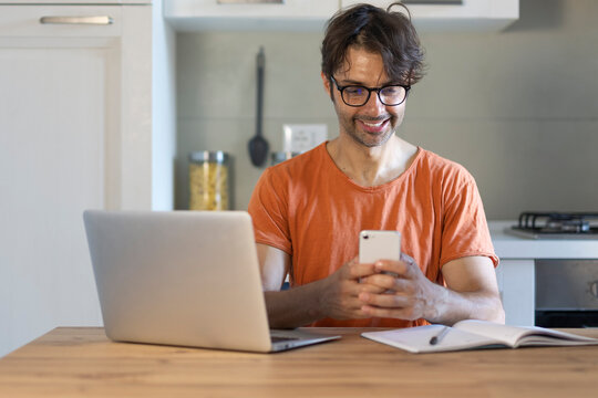 Home, cellphone and man in kitchen, laptop and online reading with creativity, project research and review. Social media, freelancer or person with pc, smartphone or feedback for proposal or chatting