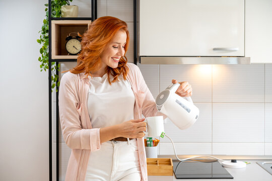 Young ginger woman pouring hot water from kettle into mug