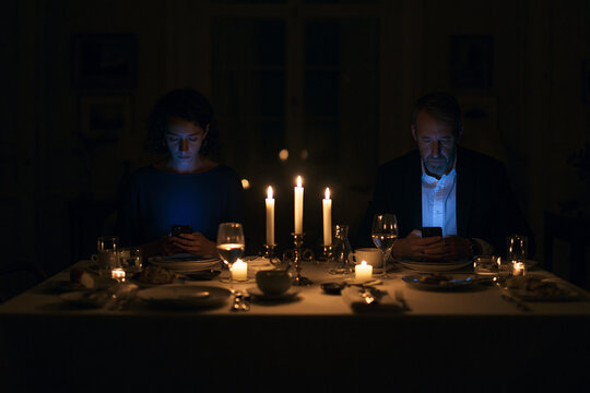 A couple at a dining table for a candlelit dinner, not interacting. Both are looking down at their own separate phones.
