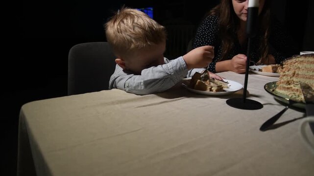 Mother watches her young son in a festive shirt and bow tie as he eagerly reaches for a piece of birthday cake while sitting at the family table during an evening celebration.