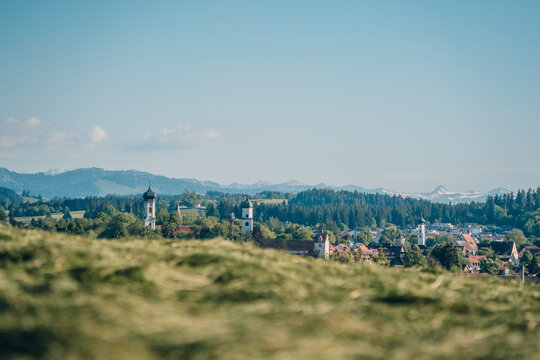 Blick &uuml;ber die Stadt Isny im Allg&auml;u mit sanften H&uuml;geln und Alpenpanorama im Hintergrund an einem sonnigen Tag.