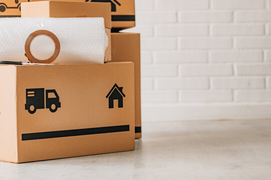 Stack of cardboard moving boxes with truck and house icons, bubble wrap, and packing tape on top, against a white brick wall. Perfect for relocation and moving concepts