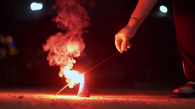 Close-up of a person using a sparkler to light an anar fountain firework during Diwali celebrations at night, with glowing flames and smoke with background of a city street