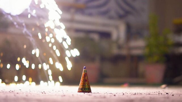 Close-up of a person using a sparkler to light an anar fountain firework during Diwali celebrations at night, with glowing flames and smoke with blurred background of a house