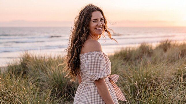 Joyful woman with wavy hair laughing over shoulder on beach at golden sunset