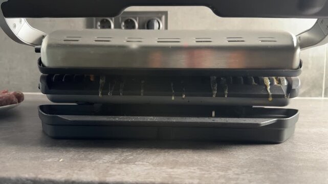Hot grease and meat juices dripping down from the plates of an electric contact grill into a black plastic drip pan on a kitchen countertop