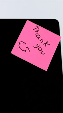A hand sticks a pink memo paper saying thank you directly onto a computer monitor screen, symbolizing office support, kindness and teamwork today.