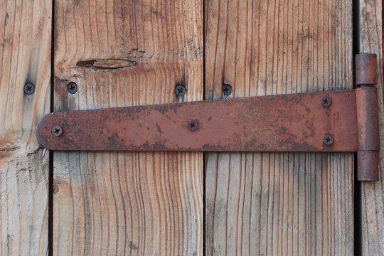 Close-up of an old, rusty metal strap hinge screwed into vertical weathered wooden planks. Rustic background detailing industrial decay, texture of aged timber, and vintage hardware.