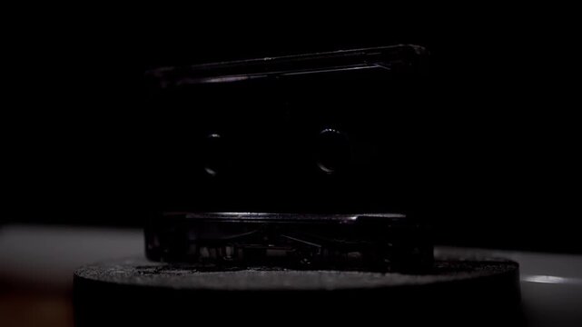 A clear audio cassette tape rotates slowly on a stand, illuminated by dramatic backlighting. Concept of vintage music technology, analog nostalgia, and the revival of classic retro media.