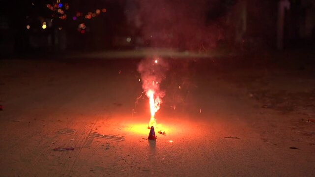 Close-up of a person using a sparkler to light an anar fountain firework during Diwali celebrations at night, with glowing flames and smoke.