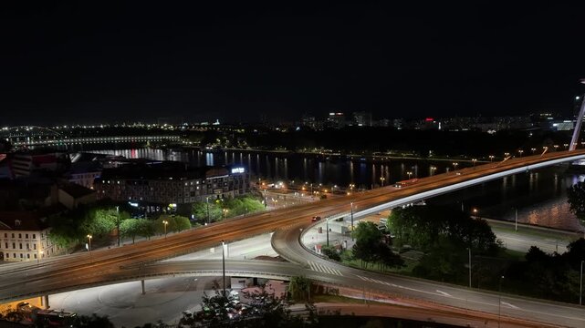 Night panorama of Bratislava with the iconic SNP Bridge illuminated above the Danube. A peaceful cityscape viewed from Bratislava Castle, with occasional cars and buses crossing the bridge