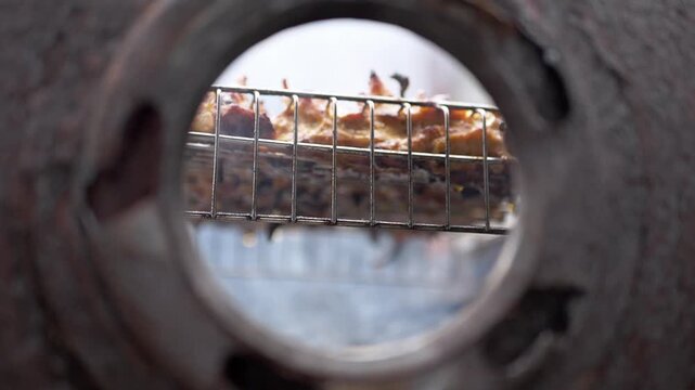 Succulent meat rotates in a grill cage seen through a circular metal frame with shallow focus. Concept of rustic food preparation, traditional grilling, and outdoor cooking.