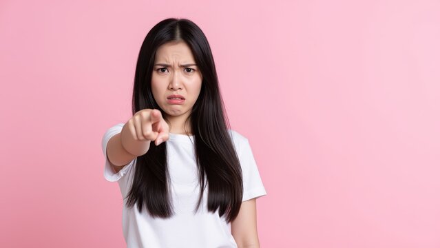 Displeased Gesture: A young Asian woman with long, dark hair frowns and points her finger accusingly against a vibrant pink backdrop, conveying disapproval.
