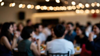 Fototapeta premium Blurred Gathering: A lively, out-of-focus scene of diverse people gathered around tables, illuminated by warm string lights, suggests a festive social event or a communal meal.