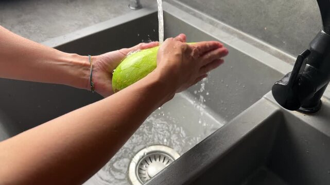 Close-up of a person's hands washing a fresh organic green zucchini under a stream of running water from the tap in a modern grey composite kitchen sink, preparing raw vegetables for a healthy meal